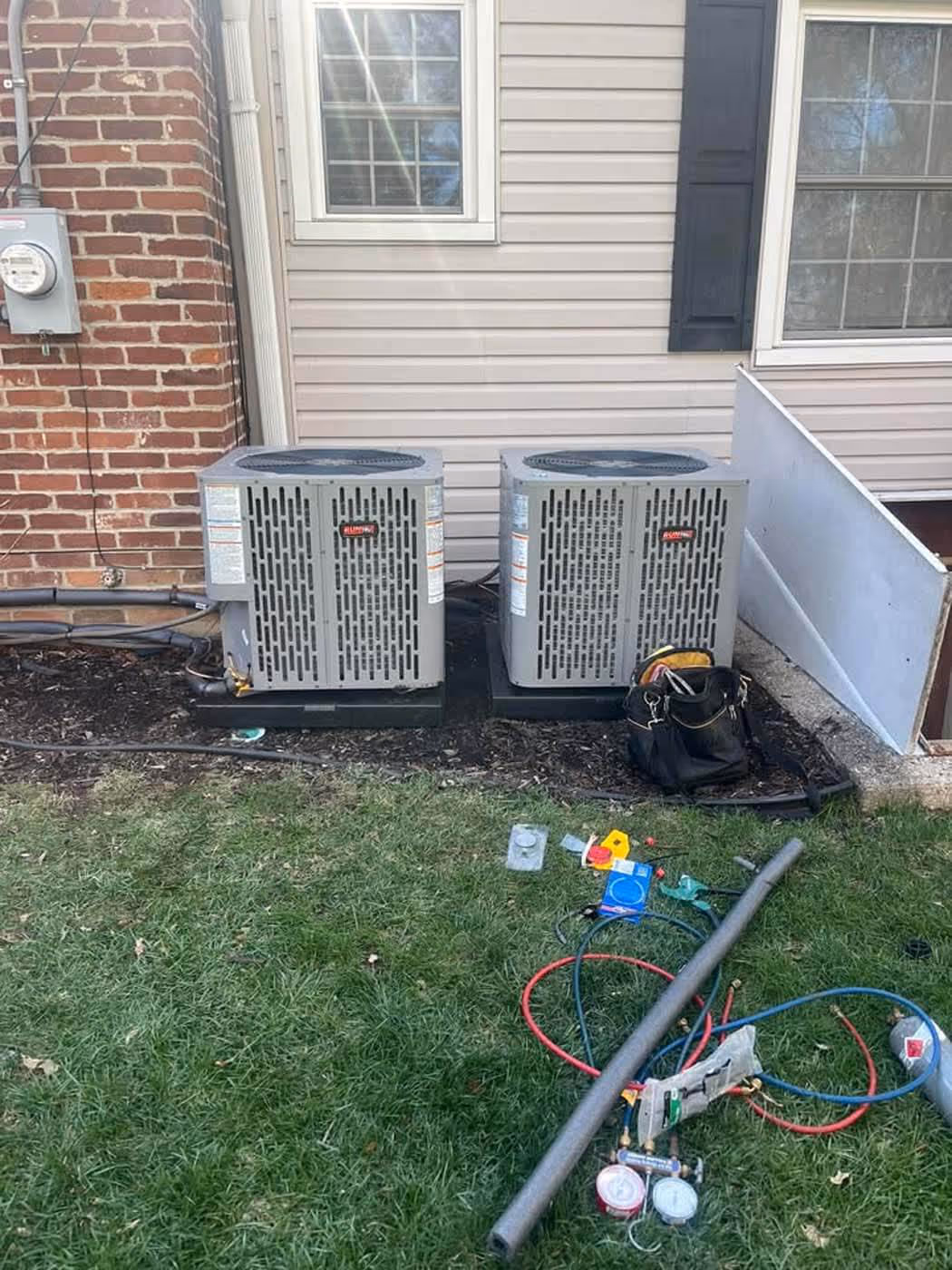 Two outdoor air conditioning units installed beside a house with tools and equipment on the grass nearby.
